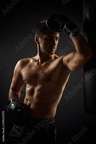 Young muscular handsome male boxer in black gloves standing near punching bag on dark background © producer