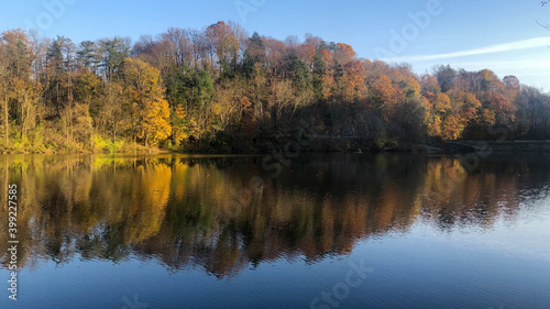Majestic fall colors line the shore and are reflected in the lake with a natural arrow pointing to a pathway in the woods. Beautiful serene water surface, foliage blazes with golden hour sunlight.