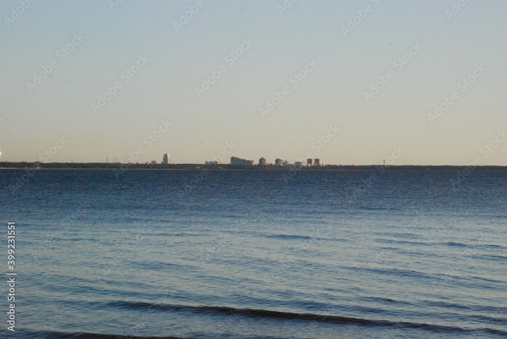 Thin coastline in the distance.
Autumn evening the sun is approaching the horizon. Blue water of the Gulf of Finland, small waves on the water. In the distance, the outlines of buildings on the shore 
