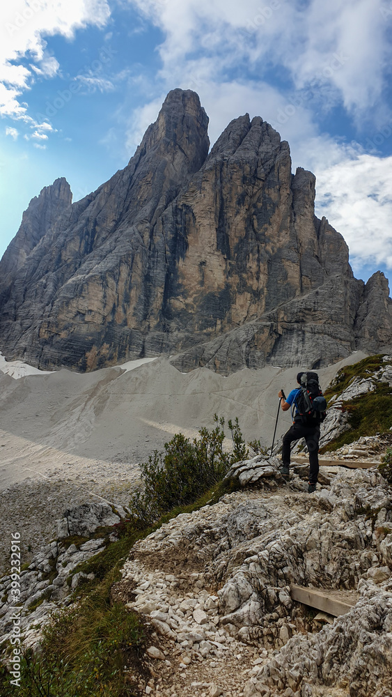 A man with big backpack hiking in high, Italian Dolomites. There are many sharp peaks behind. He is standing on a big boulder, enjoying the view. There are a few trees around. Sunny day. Outdoor