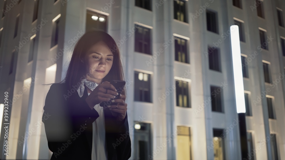 Woman at night with phone wearing black jacket and neck scarf, on background of blue buildings. Middle shot of young woman alone texting in the phone, night shot