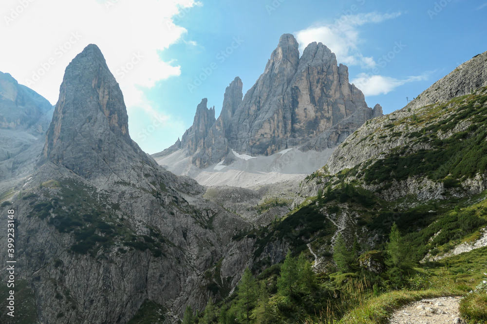 A panoramic view on Italian Dolomites. There are many high and sharp peak in front, with many landslides. Dangerous climbing. There are few trees and bushes on the side. Raw and unspoiled landscape