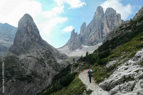 A woman with big backpack and sticks, hiking in high Italian Dolomites. There are many sharp peaks in front of her. She is going up. There are a few trees around. Sunny day. Outdoor exercising