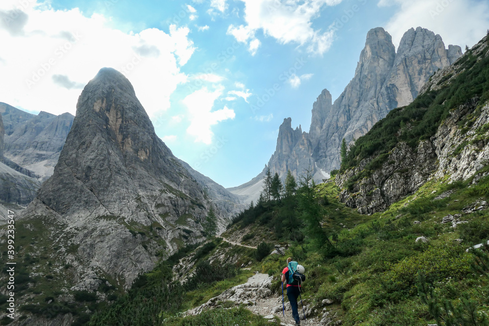 A woman with big backpack and sticks, hiking in high Italian Dolomites. There are many sharp peaks in front of her. She is going up. There are a few trees around. Sunny day. Outdoor exercising