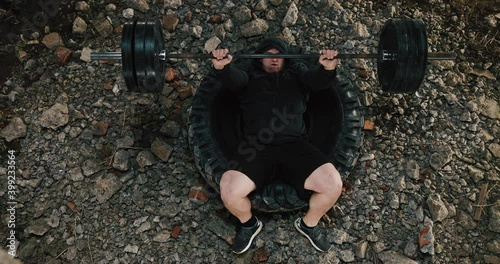 Look from top at man lifiting heavy barrel lying on the tire in an old factory. Sport, 4k photo