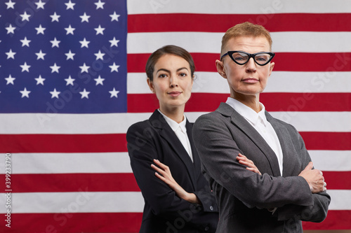 Waist up portrait of two confident female politicians looking at camera while standing with arms crossed against USA flag background, copy space photo