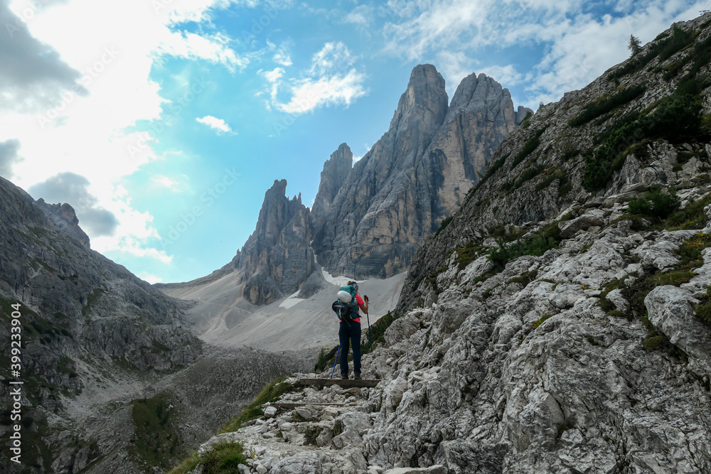 A woman with big backpack and sticks, hiking in high Italian Dolomites. There are many sharp peaks in front of her. She is going up. Lots of lose stones and landslides. Sunny day. Outdoor exercising