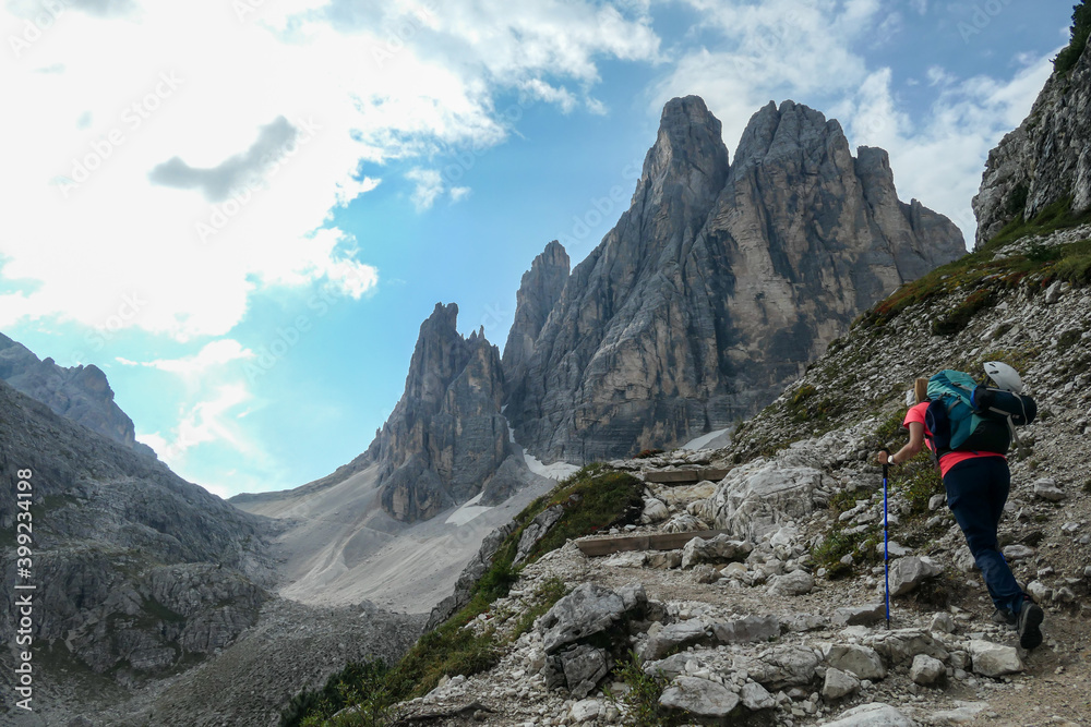 A woman with big backpack and sticks, hiking in high Italian Dolomites. There are many sharp peaks in front of her. She is going up. Lots of lose stones and landslides. Sunny day. Outdoor exercising