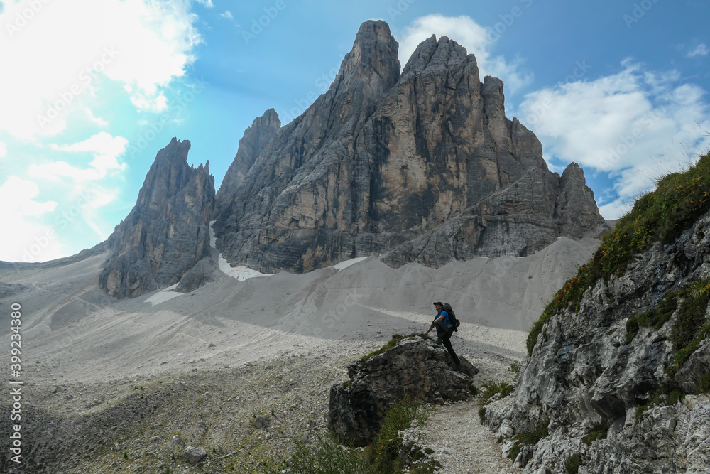 Man with big backpack and sticks, hiking in high Italian Dolomites. There are many sharp peaks in front of him. He is standing on a big boulder. Lots of lose stones and landslides. Sunny day. Outdoor