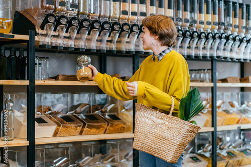 Fotografie Minimalist vegan style girl with wicker bag and reusable glass coffee cup on background of interior of zero waste shop