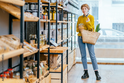 Minimalist vegan style girl with wicker bag and reusable glass coffee cup on background of interior of zero waste shop. Woman doing shopping without plastic packaging in plastic free grocery store. photo