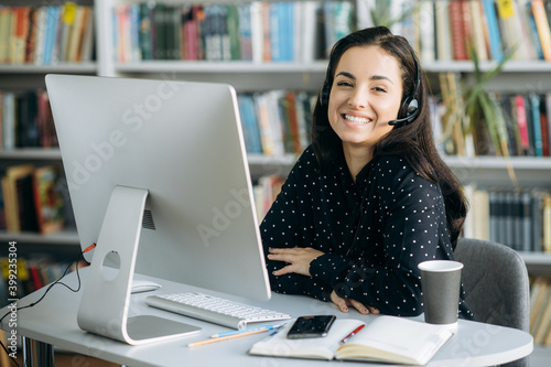 Remote work. Happy young pretty woman, call center worker, teacher or manager, wearing a headset, looks at the camera with smile while sitting at her workplace