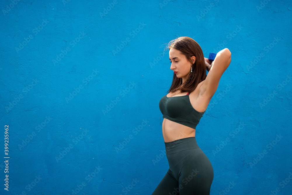 Fototapeta premium Pretty young woman in green sportswear holds dumbbells in her hands over on blue background