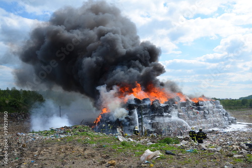 A group of firefighters extinguishes huge landfill fire with flames and black smoke in the background