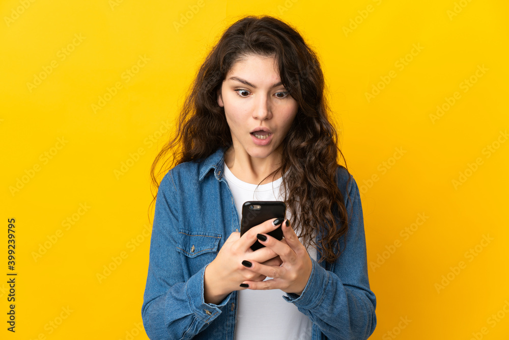 Teenager Russian girl isolated on yellow background looking at the camera while using the mobile with surprised expression Teenager Russian girl isolated on yellow background looking at the camera while using the mobile with surprised expression