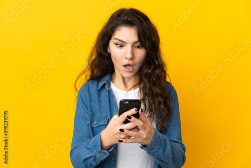 Teenager Russian girl isolated on yellow background looking at the camera while using the mobile with surprised expression