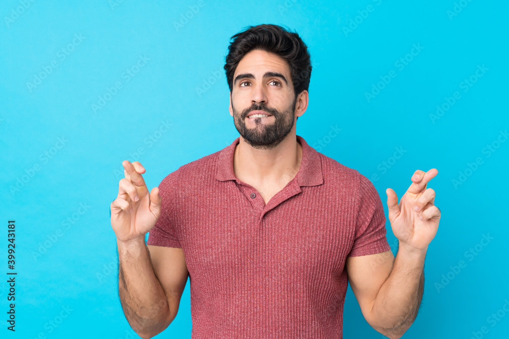 Young handsome man with beard over isolated blue background with fingers crossing and wishing the best Young handsome man with beard over isolated blue background with fingers crossing and wishing the best