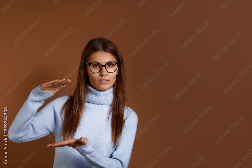 Young caucasian woman wearing glasses and light blue sweater shows big size of present, shapes height of gift box with hands, shapes huge box, demonstrates increase, isolated over brown background. Young caucasian woman wearing glasses and light blue sweater shows big size of present, shapes height of gift box with hands, shapes huge box, demonstrates increase, isolated over brown background.