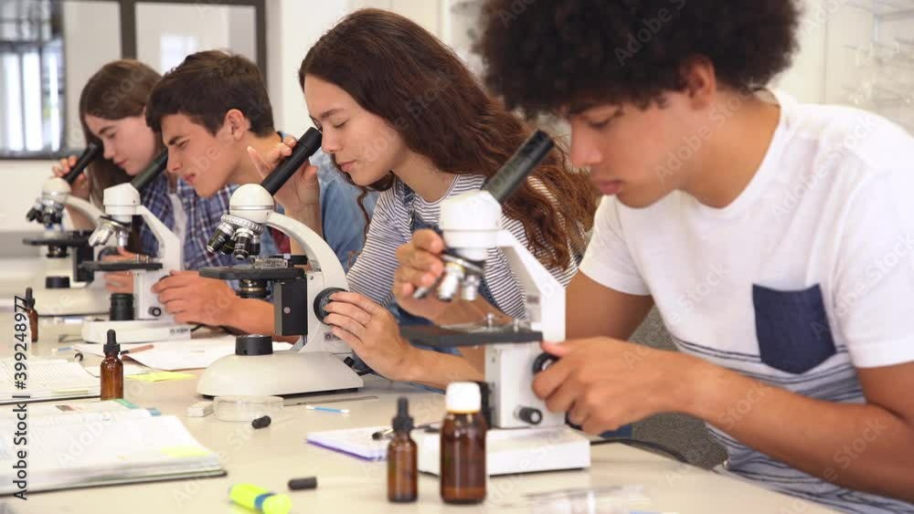 Group of multiethnic college students performing experiment using microscope in science lab. University focused students looking through microscope in biology class while sitting in a row at desk.