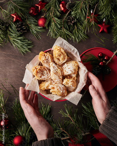 Round red box with pastry on dark brown wooden table, young woman’s hands holding it from the bottom right. Pine branches and cones laying on the table, with red Christmas decorations photo