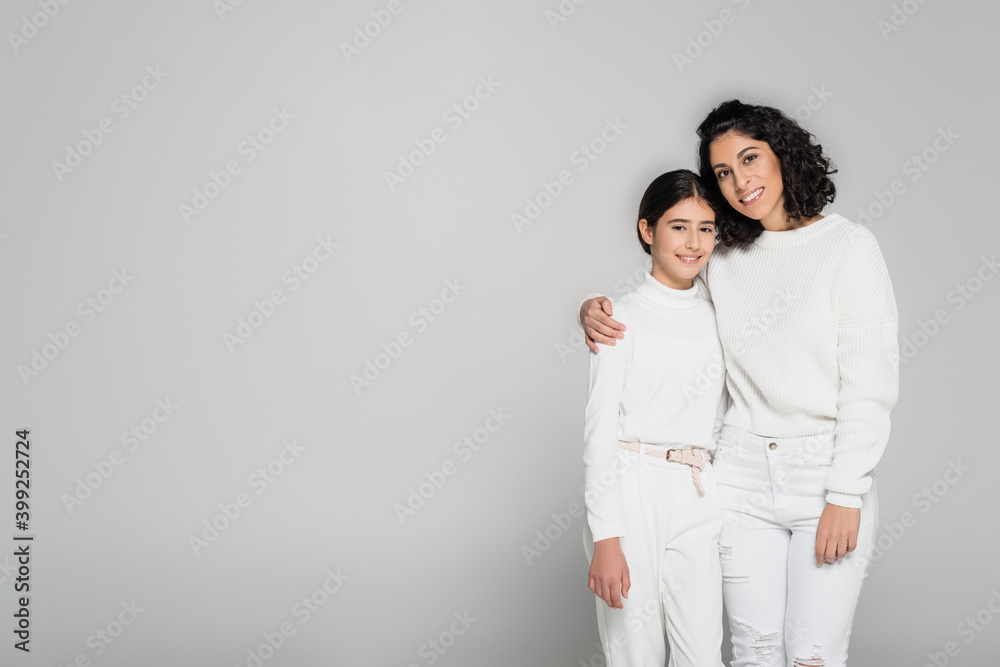 Cheerful hispanic woman hugging child and looking at camera on grey background, two generations of women Cheerful hispanic woman hugging child and looking at camera on grey background, two generations of women