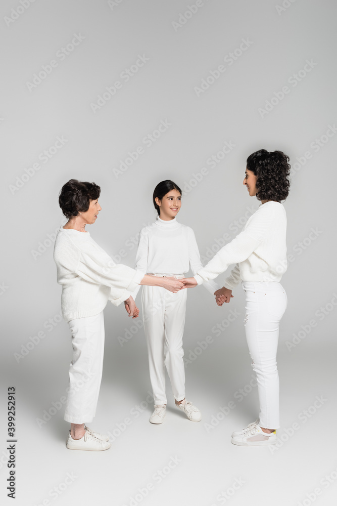 Three generations of hispanic women holding hands and smiling at each other on grey background