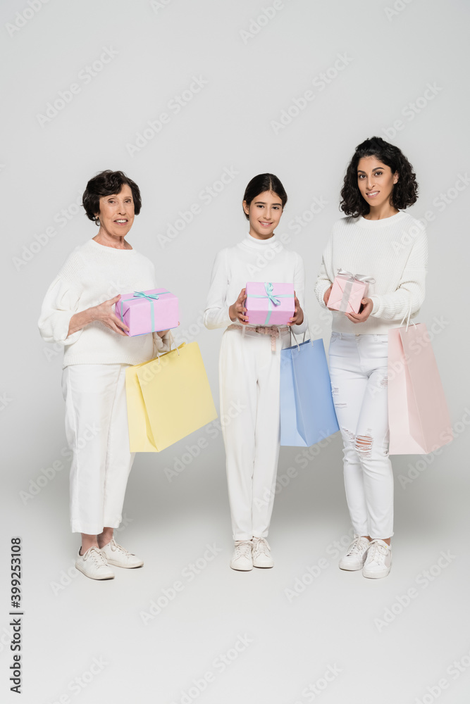 Smiling hispanic women and girl holding shopping bags and gifts on grey background, three generations of women Smiling hispanic women and girl holding shopping bags and gifts on grey background, three generations of women
