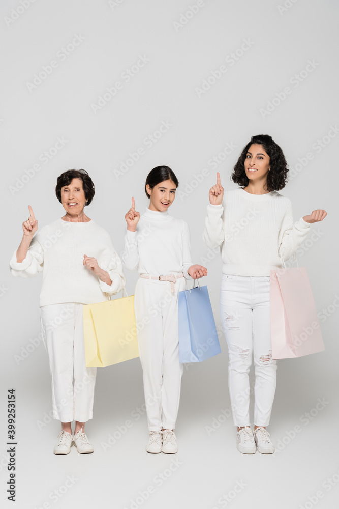 Cheerful hispanic women and girl pointing with fingers and holding shopping bags on grey background, three generations of women Cheerful hispanic women and girl pointing with fingers and holding shopping bags on grey background, three generations of women