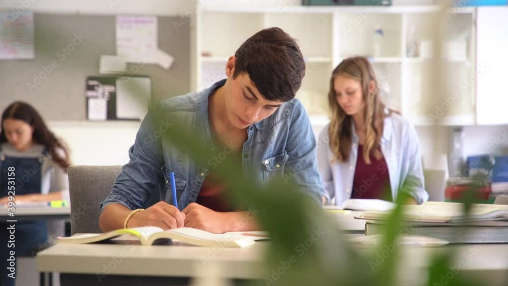 Happy student taking notes while studying in high school. Satisfied young man looking at camera while sitting at desk in classroom. Portrait of college guy writing while completing project. Happy student taking notes while studying in high school. Satisfied young man looking at camera while sitting at desk in classroom. Portrait of college guy writing while completing project.