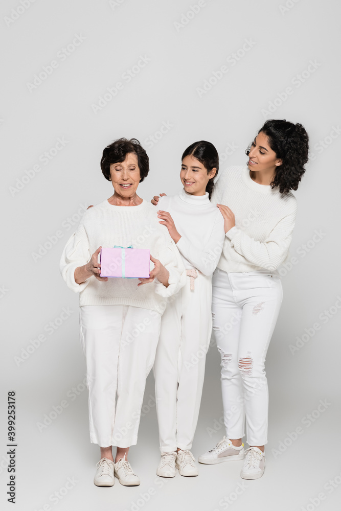 Smiling hispanic woman and daughter embracing granny with present on grey background, three generations of women