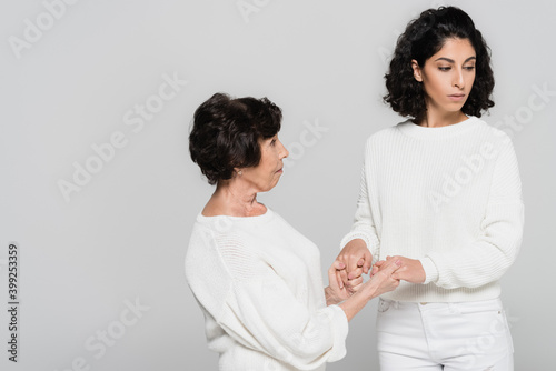 Hispanic senior woman holding hands of sad daughter isolated on grey, two generations of women