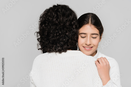 Positive hispanic girl hugging mother with closed eyes isolated on grey  two generations of women