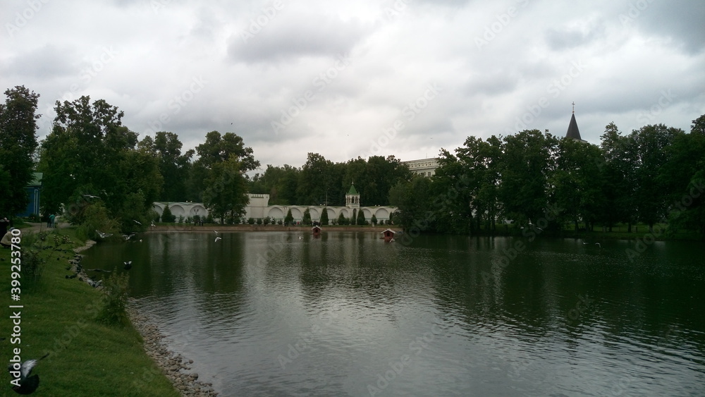 The gray surface of the lake with reflected clouds and white buildings hidden behind green trees The gray surface of the lake with reflected clouds and white buildings hidden behind green trees