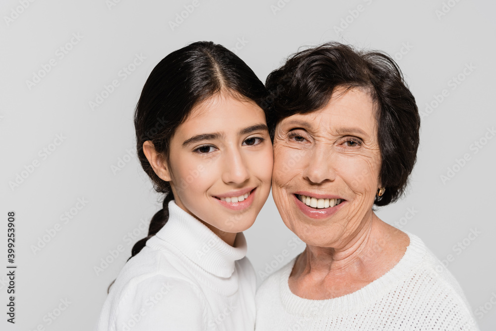Senior woman smiling near hispanic granddaughter isolated on grey, two generations of women Senior woman smiling near hispanic granddaughter isolated on grey, two generations of women