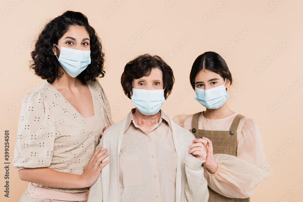 Hispanic woman and daughter hugging grandmother in medical mask isolated on beige, three generations of women