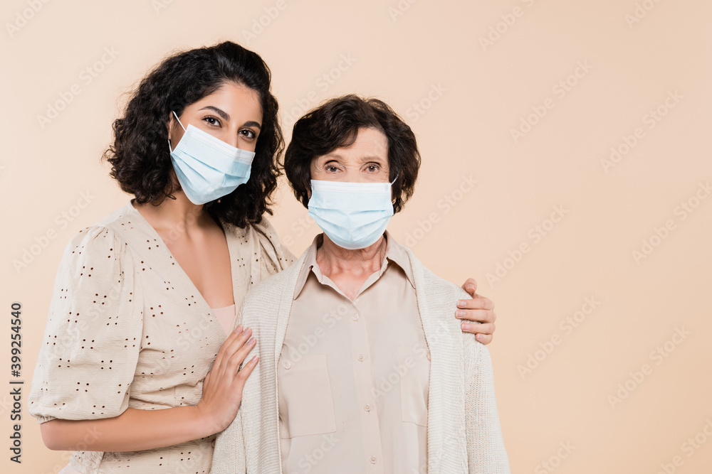 Hispanic woman embracing senior mother in medical mask isolated on beige, two generations of women