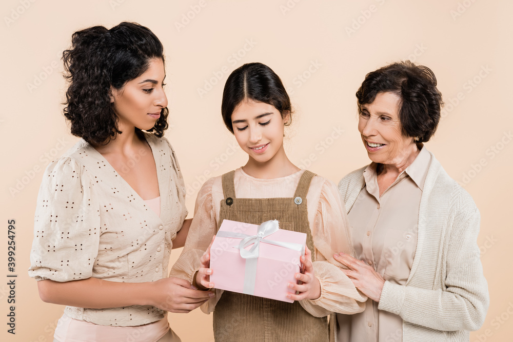 Smiling hispanic child holding gift box near mother and grandmother isolated on beige, three generations of women Smiling hispanic child holding gift box near mother and grandmother isolated on beige, three generations of women
