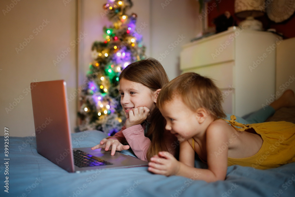 Girls children in bedroom are looking at screen near Christmas tree in real interior, concept of Christmas sales and interesting cartoons. siblings on the bed with blanket lying looking laptop Girls children in bedroom are looking at screen near Christmas tree in real interior, concept of Christmas sales and interesting cartoons. siblings on the bed with blanket lying looking laptop