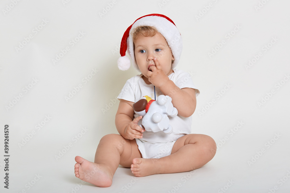 Baby boy wearing Santa Claus hat and white background, playing with plastic dog toy, looking away, biting his fingers, sitting barefoot on floor, looking away. Baby boy wearing Santa Claus hat and white background, playing with plastic dog toy, looking away, biting his fingers, sitting barefoot on floor, looking away.