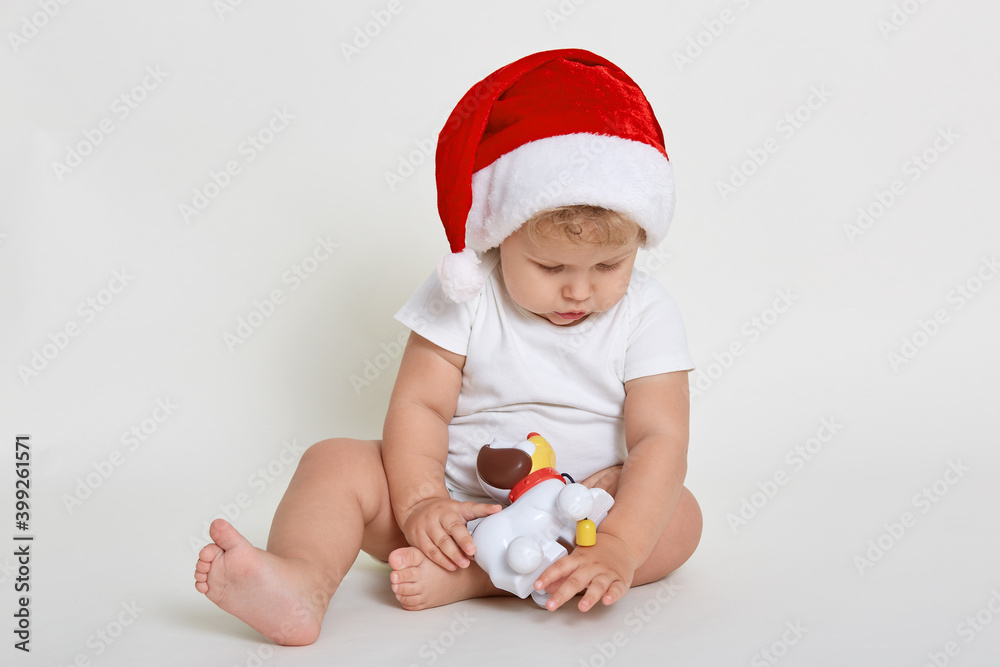 Santa boy playing with fog toy isolated over white background, barefoot infant in bodysuit looking concentrated at toy, infant looking down, cute baby on Christmas Eve. Santa boy playing with fog toy isolated over white background, barefoot infant in bodysuit looking concentrated at toy, infant looking down, cute baby on Christmas Eve.