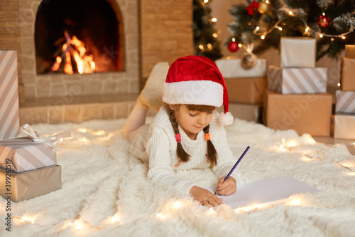 Little girl in red christmas hat writes letter to Santa Claus, adorable child lying on floor near fireplace, wearing white warm jumper, lying on floor on soft carpet. photo