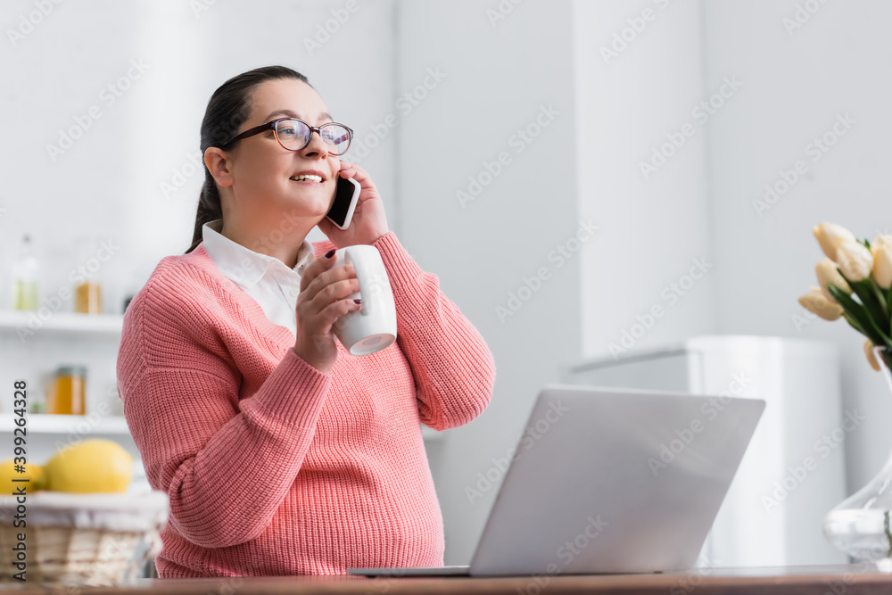 Smiling plus size hispanic woman with cup talking on smartphone in kitchen on blurred foreground Smiling plus size hispanic woman with cup talking on smartphone in kitchen on blurred foreground