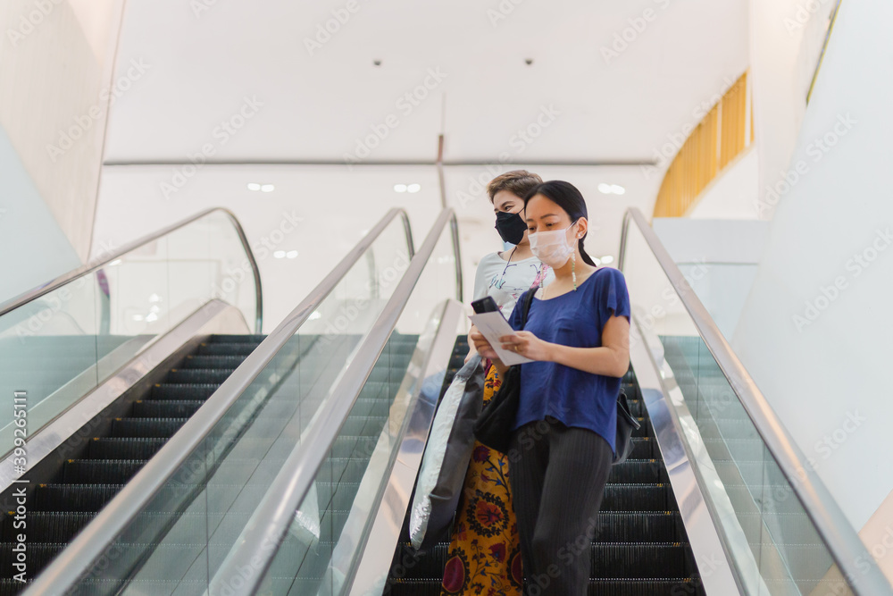 Two woman in protective mask with shopping bags and mobile phone standing on escalator in shopping mall.