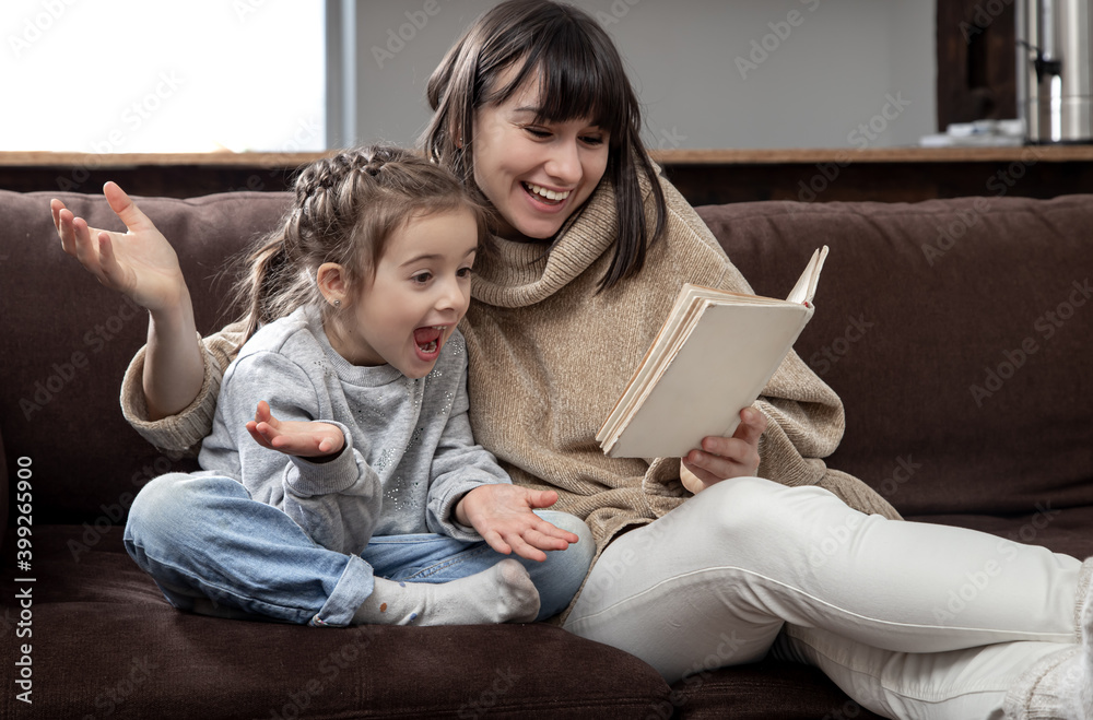 Young mother with her daughter are reading a book at home while sitting on the couch. Young mother with her daughter are reading a book at home while sitting on the couch.
