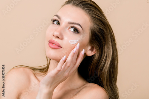 Young European blond woman with moisturizing cream on cheek. Beauty shot of beautiful woman touching her fresh glowing hydrated facial skin on beige background. Youth and Skin care concept