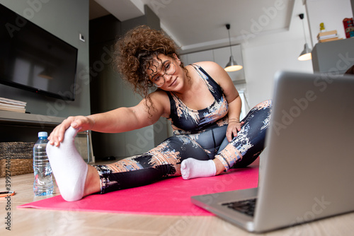 Young plus size woman at home doing exercise in front of open laptop, repeating instructions by professional online fitness trainer.
