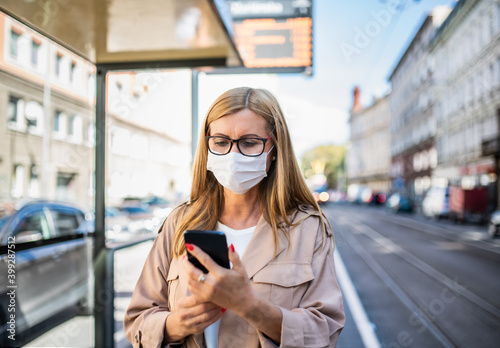 Senior woman with smartphone on bus stop outdoors in city or town, coronavirus concept.