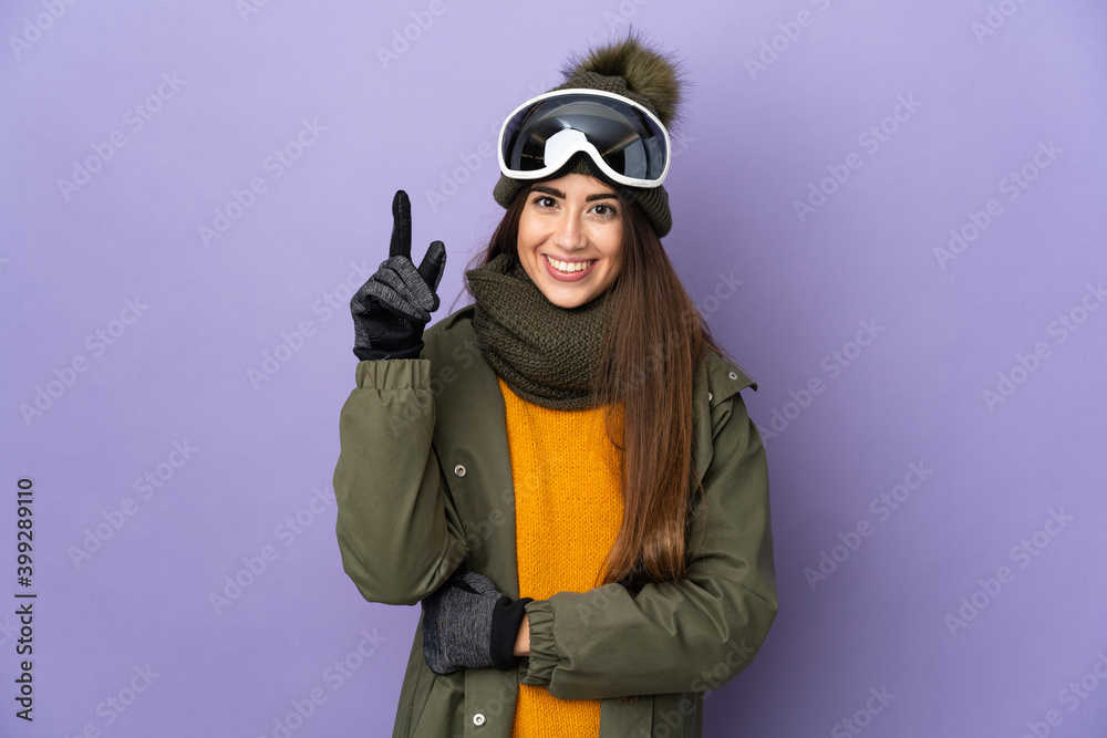 Skier caucasian girl with snowboarding glasses isolated on purple background pointing up a great idea