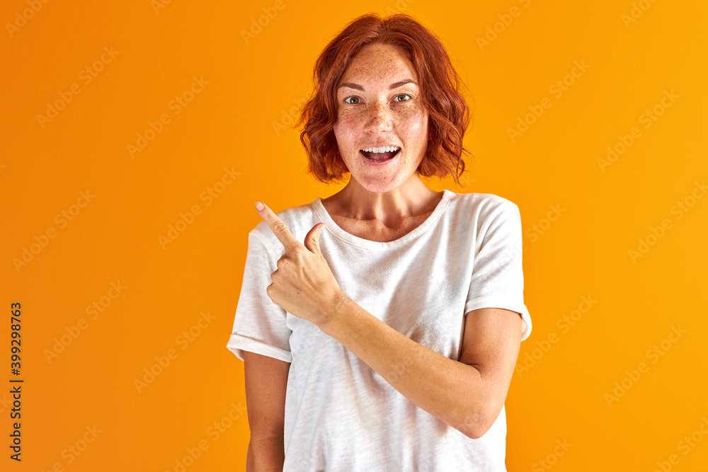 woman pointing finger at side, index, isolated over orange background. redhead female smile, in casual wear woman pointing finger at side, index, isolated over orange background. redhead female smile, in casual wear
