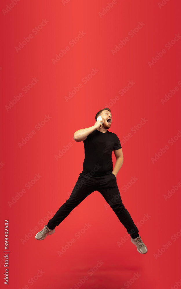 Talking phone. Full length portrait of young high jumping man gesturing isolated on red studio background. Attractive male caucasian model. Copyspace. Human emotions, facial expression concept.
